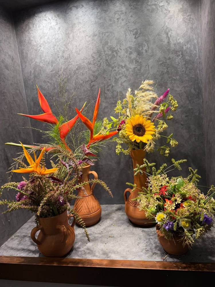 Four terracotta vases of heliconia, sunflower, celosia and grasses on a grey plaster shelf